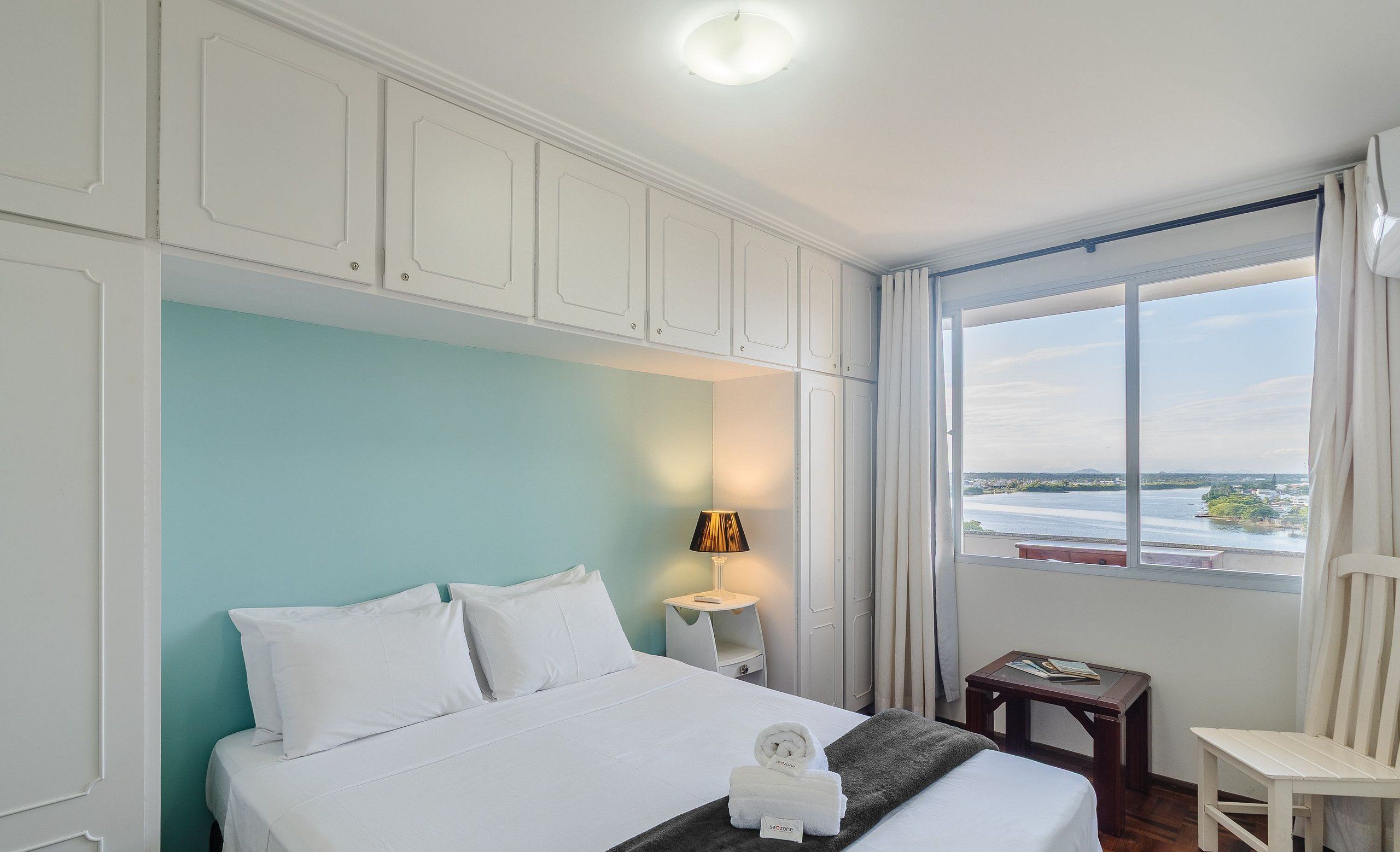 Una habitación de hotel con cama, mesita de noche con lámpara, una silla y vista al agua.