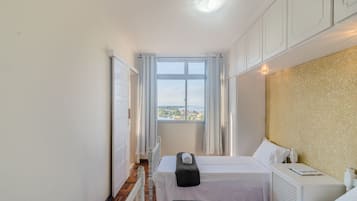A small, bright room with two beds, a window with curtains, a radiator, and a white cabinet.