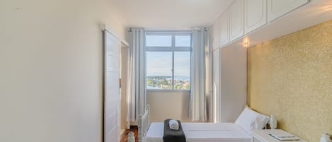 Une petite pièce lumineuse, avec deux lits, une fenêtre avec des rideaux, un radiateur et une armoire blanche.