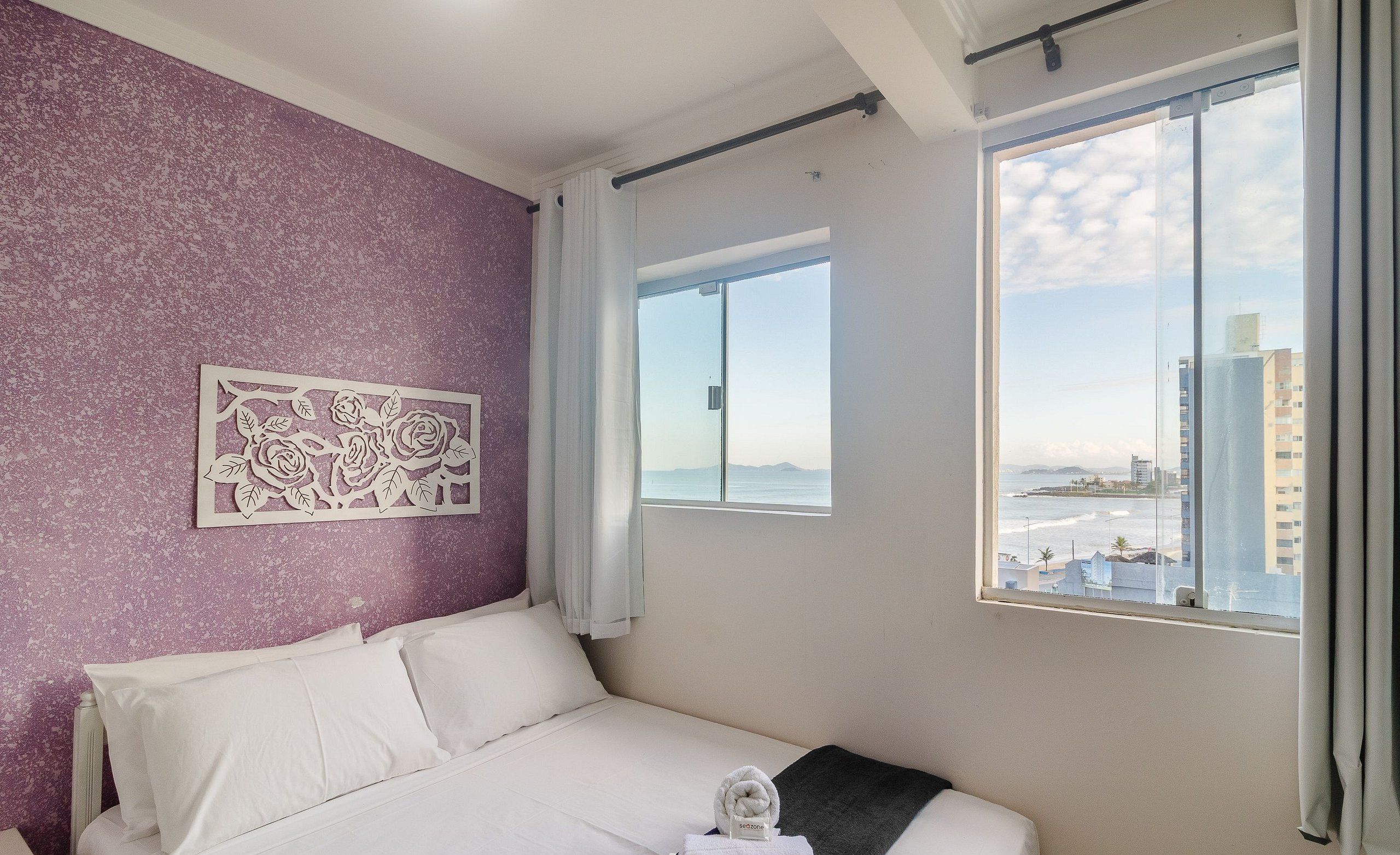 A bedroom with a bed, a window with a view of buildings and the sea, a decorative wall piece, and a towel on the bed.