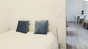 Ein Schlafzimmer mit einem Bett, zwei Kissen, einer weißen Wand, einer Tür zu einem Essbereich mit Tisch und Stühlen sowie einem Lichtschalter.