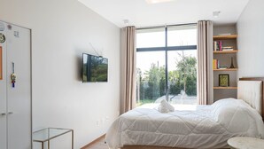 A modern bedroom with a large bed, a TV mounted on the wall, a bookshelf, and a view of greenery outside. - Comfortable House Near Praia do Rosa Egi001 (Imbituba)