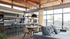 A modern kitchen with a central island, bar stools, and a dining area with a grey sofa and chairs. - Comfortable House Near Praia do Rosa Egi001 (Imbituba)