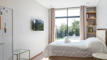 Quarto moderno com cama grande, TV fixada na parede, estante e vista para o verde lá fora.