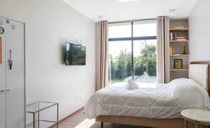 Quarto moderno com cama grande, TV fixada na parede, estante e vista para o verde lá fora.