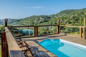 A wooden deck with a swimming pool, overlooking a hillside with houses.