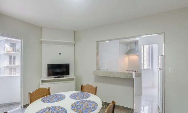 Comedor con mesa redonda, sillas, televisor y zona de cocina visible a través de una puerta abierta.