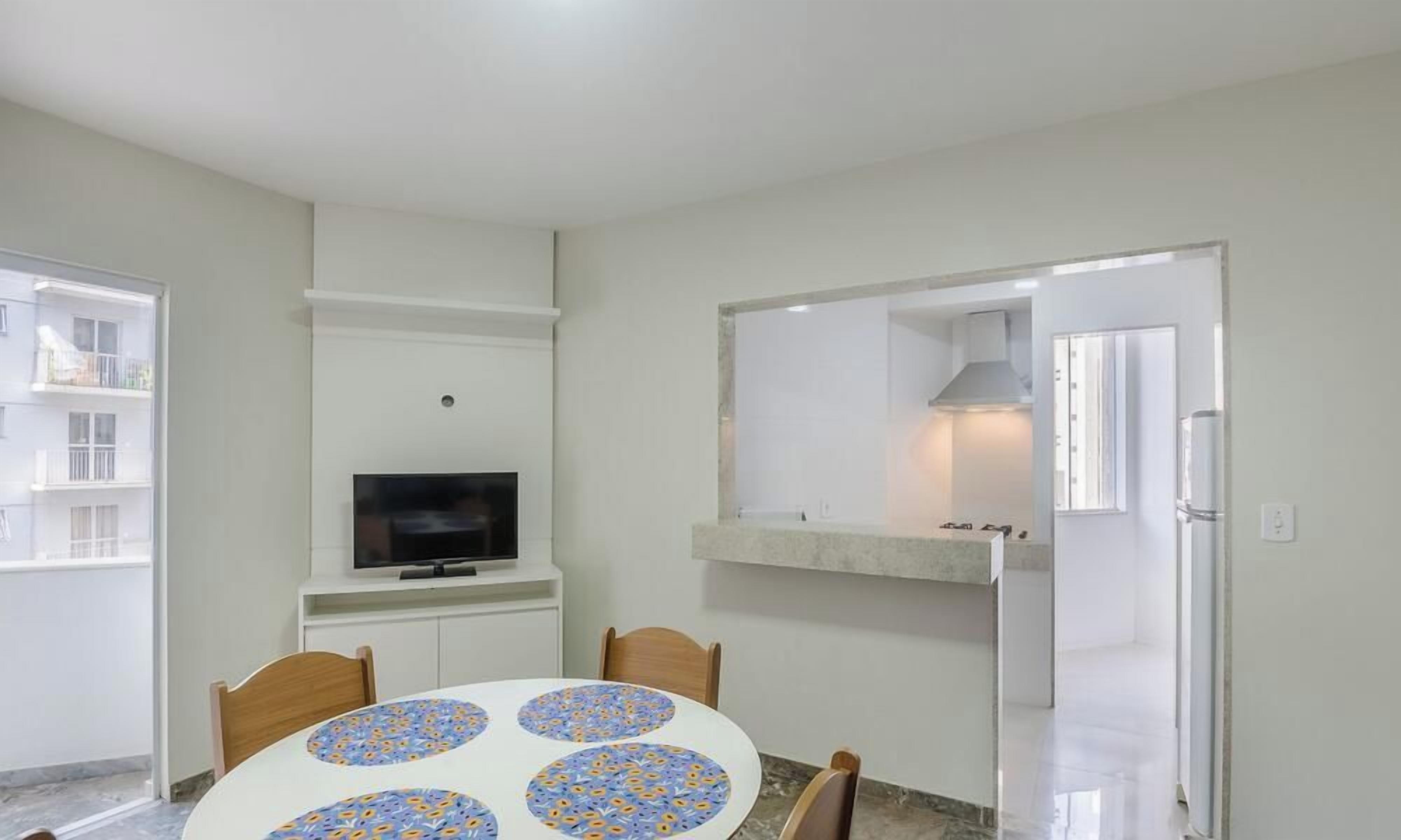 A room with a round table, chairs, a television, a kitchen area with a stove, and a door leading to another room.