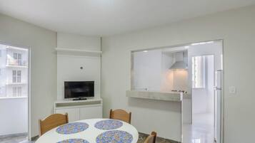 A room with a round table, chairs, a television, a kitchen area with a stove, and a door leading to another room.