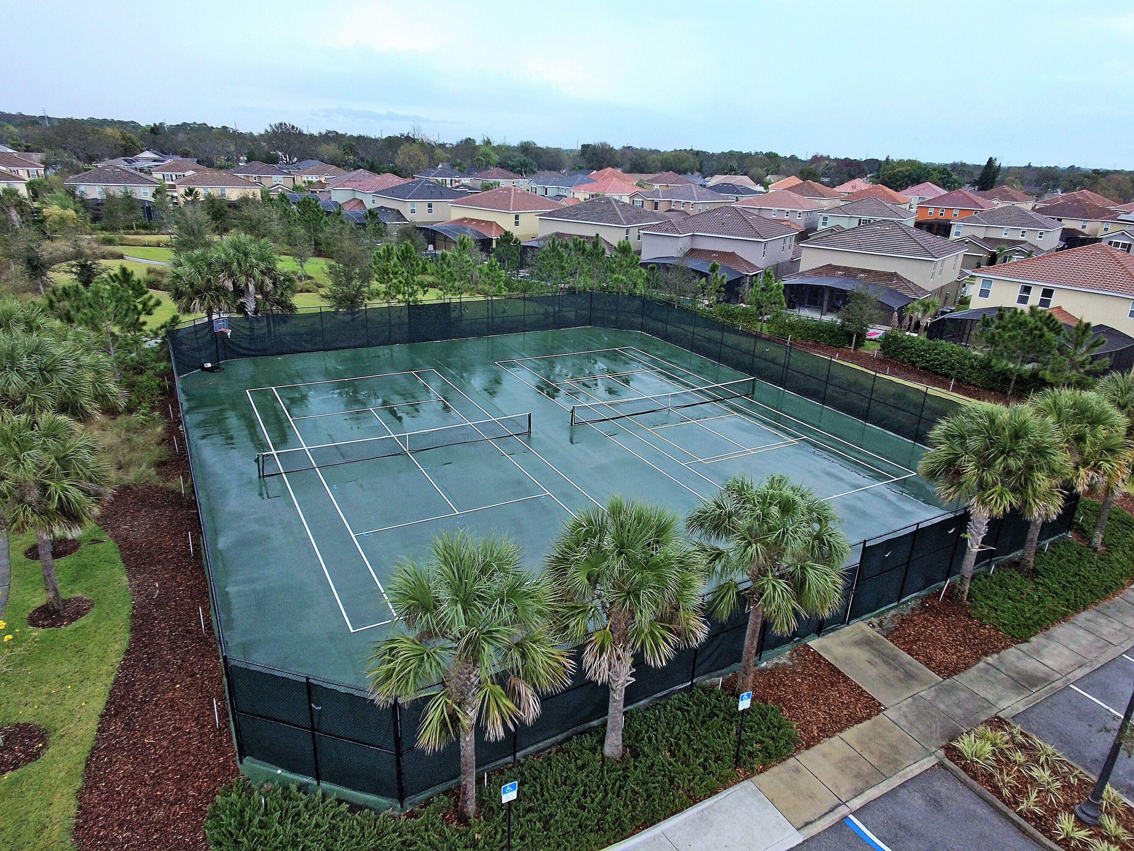 Luchtbeeld van een tennisbaan, omringd door woonhuizen en palmbomen.