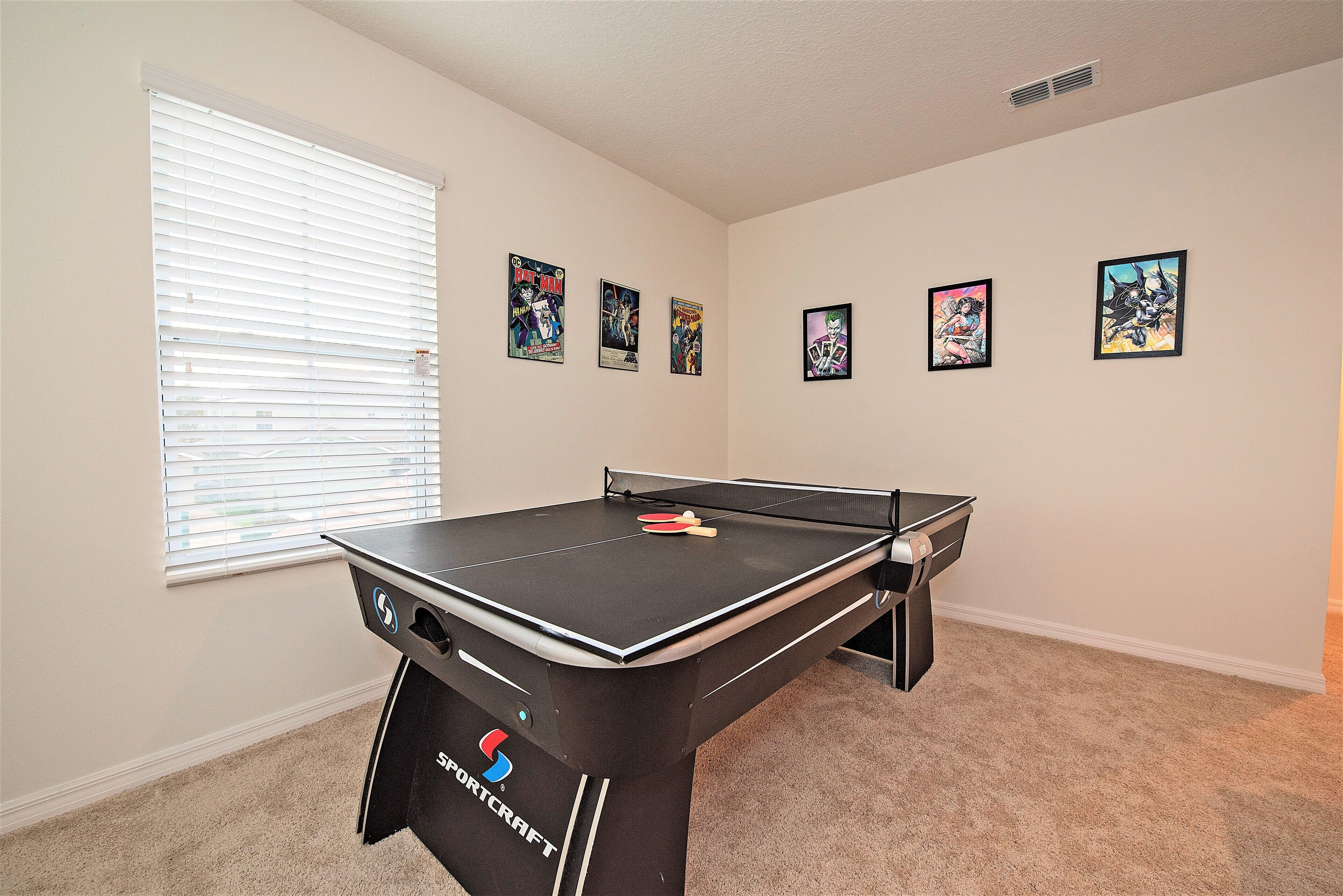 A room with a ping pong table, a window with blinds, and four framed comic book posters on the wall.