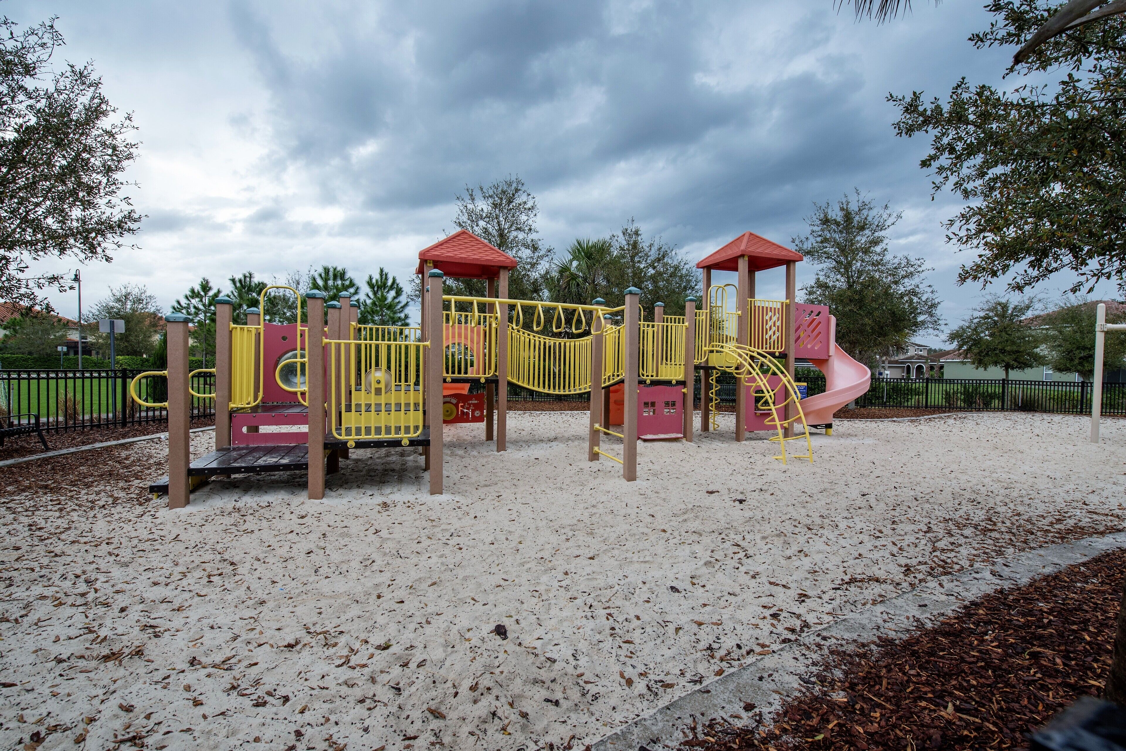 A playground with yellow and pink play structures, including a slide and climbing elements, set on a sandy surface with trees in the background.