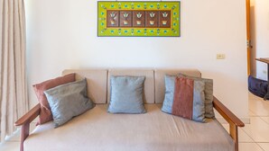 A beige sofa with three cushions in different colors, a decorative wall piece above it, and a wooden chair to the right. - Beautiful Apart With Balcony and Barbecue Ecu102 (Ubatuba)