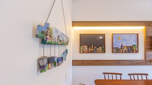A dining area with a wooden table and chairs, a wall-mounted art piece depicting a cityscape, and a hanging light fixture. - Beautiful Apart With Balcony and Barbecue Ecu102 (Ubatuba)