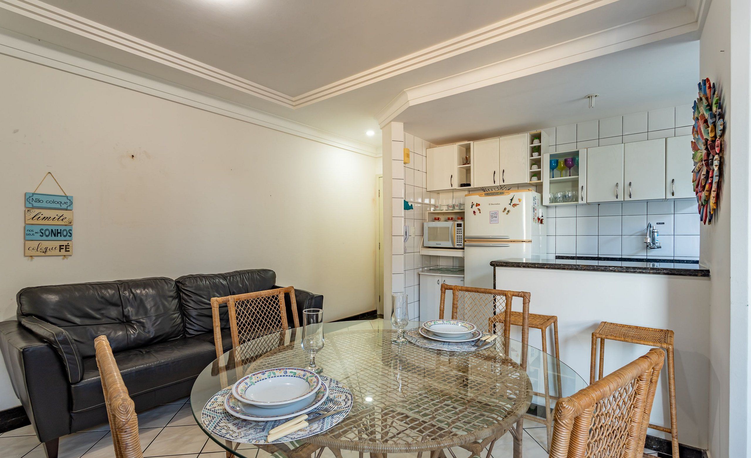 A living room with a glass-top dining table, black leather sofa, wicker chairs, and a kitchen with white cabinets and a microwave.