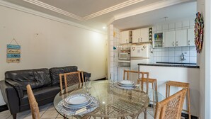 Un salon comprenant une table à manger avec un plateau en verre, un canapé en cuir noir, des fauteuils en osier et une cuisine avec des meubles blancs et un four à micro-ondes.