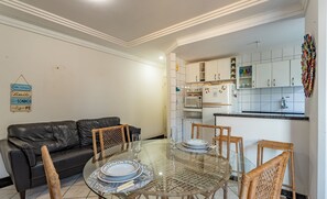 Un salon comprenant une table à manger avec un plateau en verre, un canapé en cuir noir, des fauteuils en osier et une cuisine avec des meubles blancs et un four à micro-ondes.