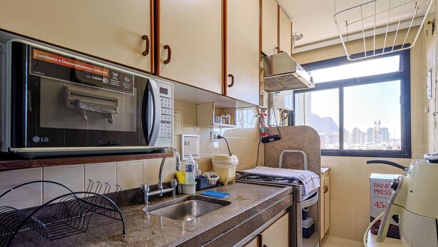 A compact kitchen with a microwave, stove, and sink. There is a window with a view of buildings. - Barra da Tijuca Vista Mar Lazer e Garagem (Rio de Janeiro)