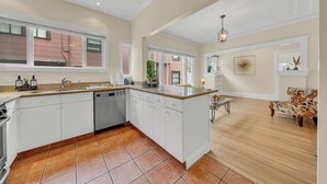 A spacious kitchen with white cabinets, a dishwasher, and a large island. There is a dining area with a patterned sofa and a chandelier.