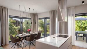 Une cuisine moderne dotée d’un plan de travail en marbre, d’un coin repas avec une table dressée pour quatre personnes et de grandes fenêtres offrant une vue sur un jardin.