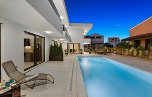 Un espace piscine extérieur moderne, doté de chaises longues, d’un coin salon abrité et d’une vue sur les maisons voisines.