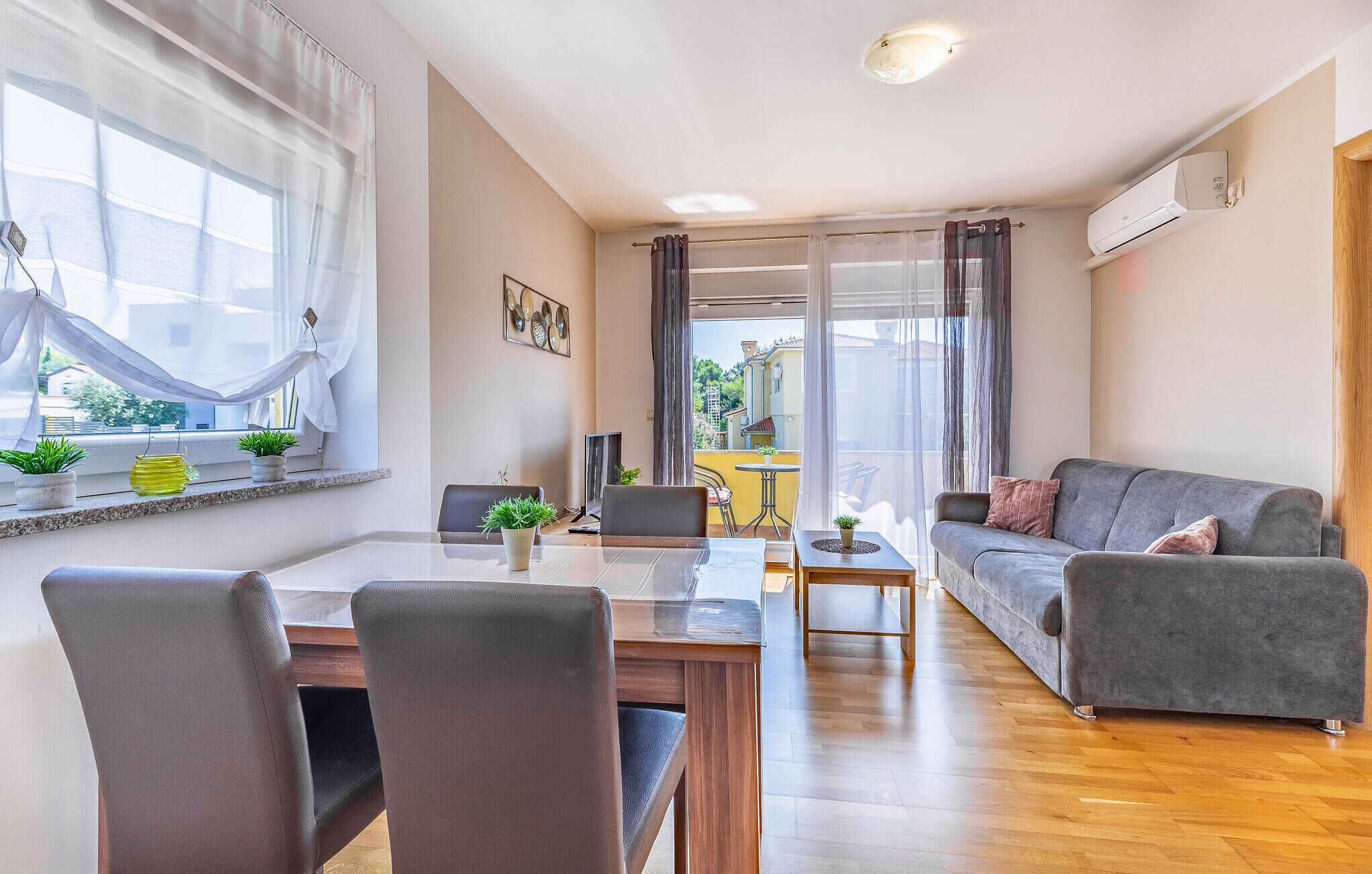 En moderne stue med en grå sofa, et spisebord med stole, en balkon med udsigt til bygninger, og et vindue med lette gardiner.