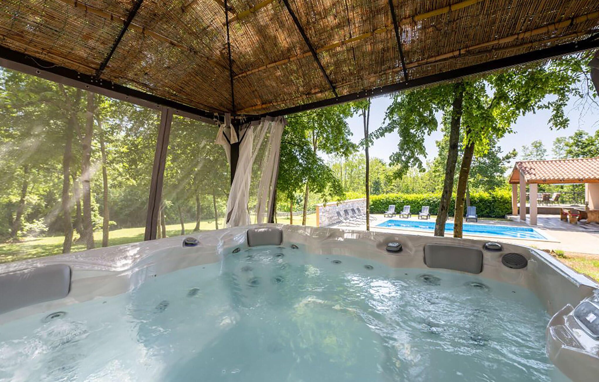 Ein Bereich mit Whirlpool, überdacht mit einer Bambuskonstruktion, umgeben von Bäumen und einem Pool im Hintergrund.