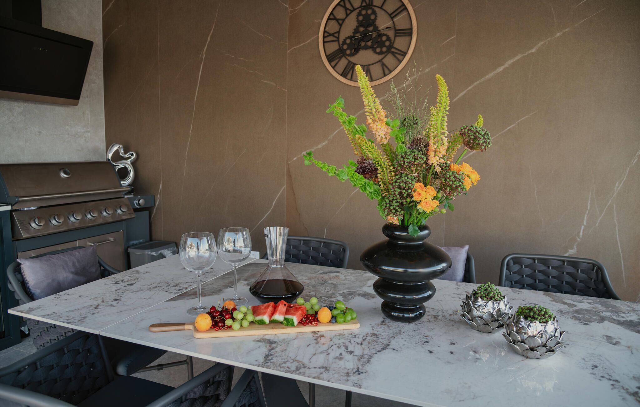 Une table à manger avec un plateau en marbre, entourée de chaises, un vase rempli de fleurs, une corbeille de fruits et une carafe à vin.