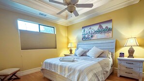 Un dormitorio con una cama, dos mesitas de noche, un ventilador de techo, una ventana con persianas y un cuadro en la pared.