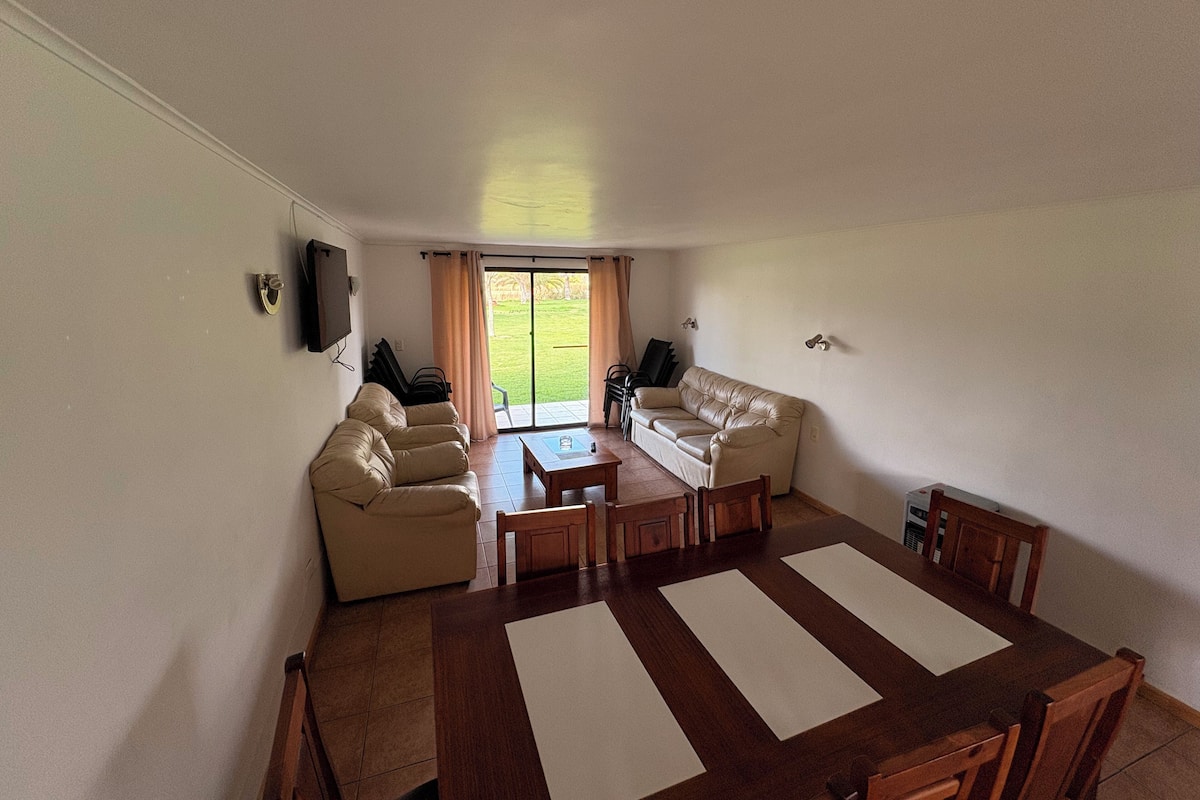 A spacious living room with a large wooden dining table, two beige sofas, and a television mounted on the wall.