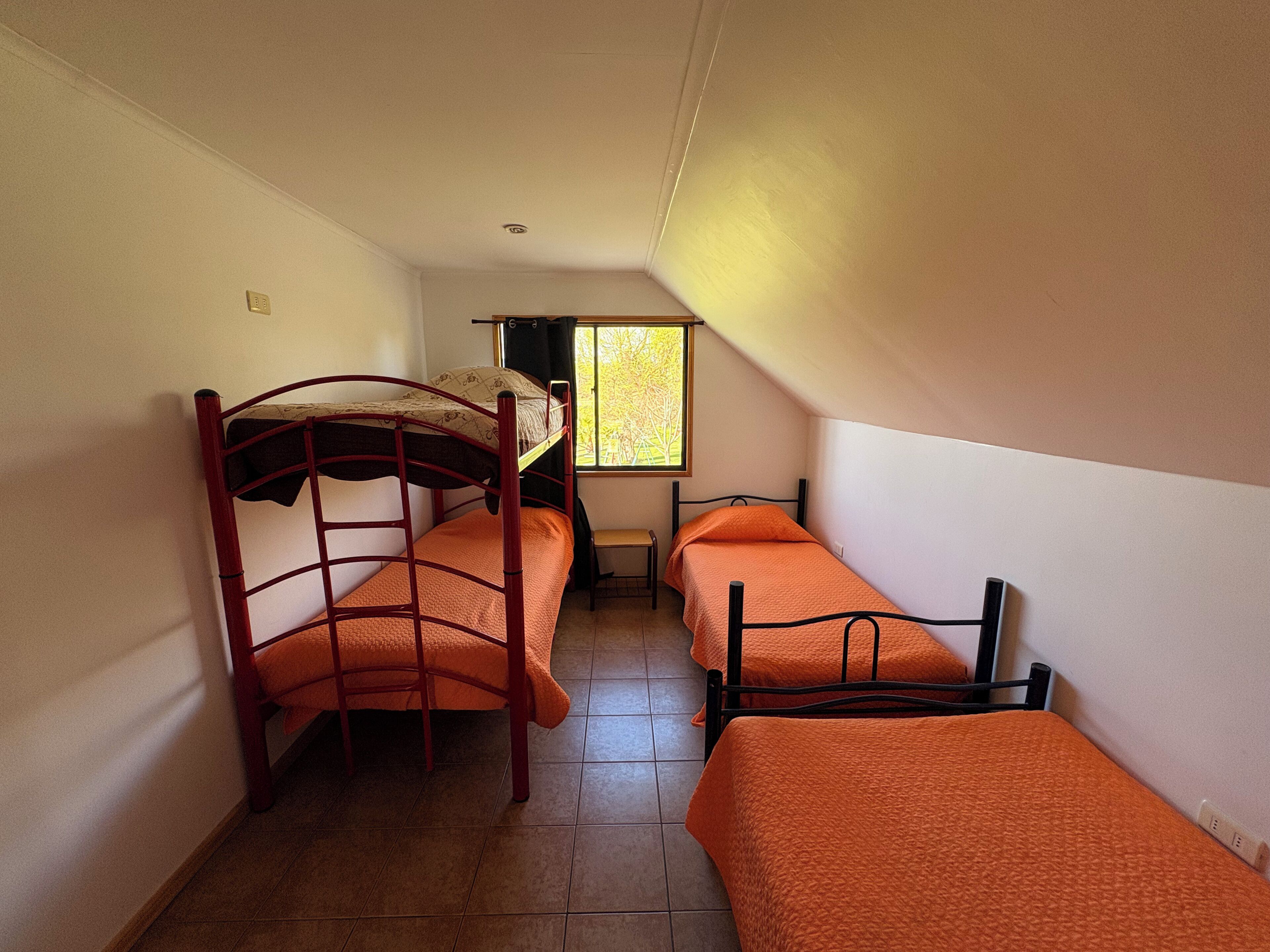Un dormitorio con dos literas y una cama individual, una ventana con vistas a árboles y suelo de baldosas.