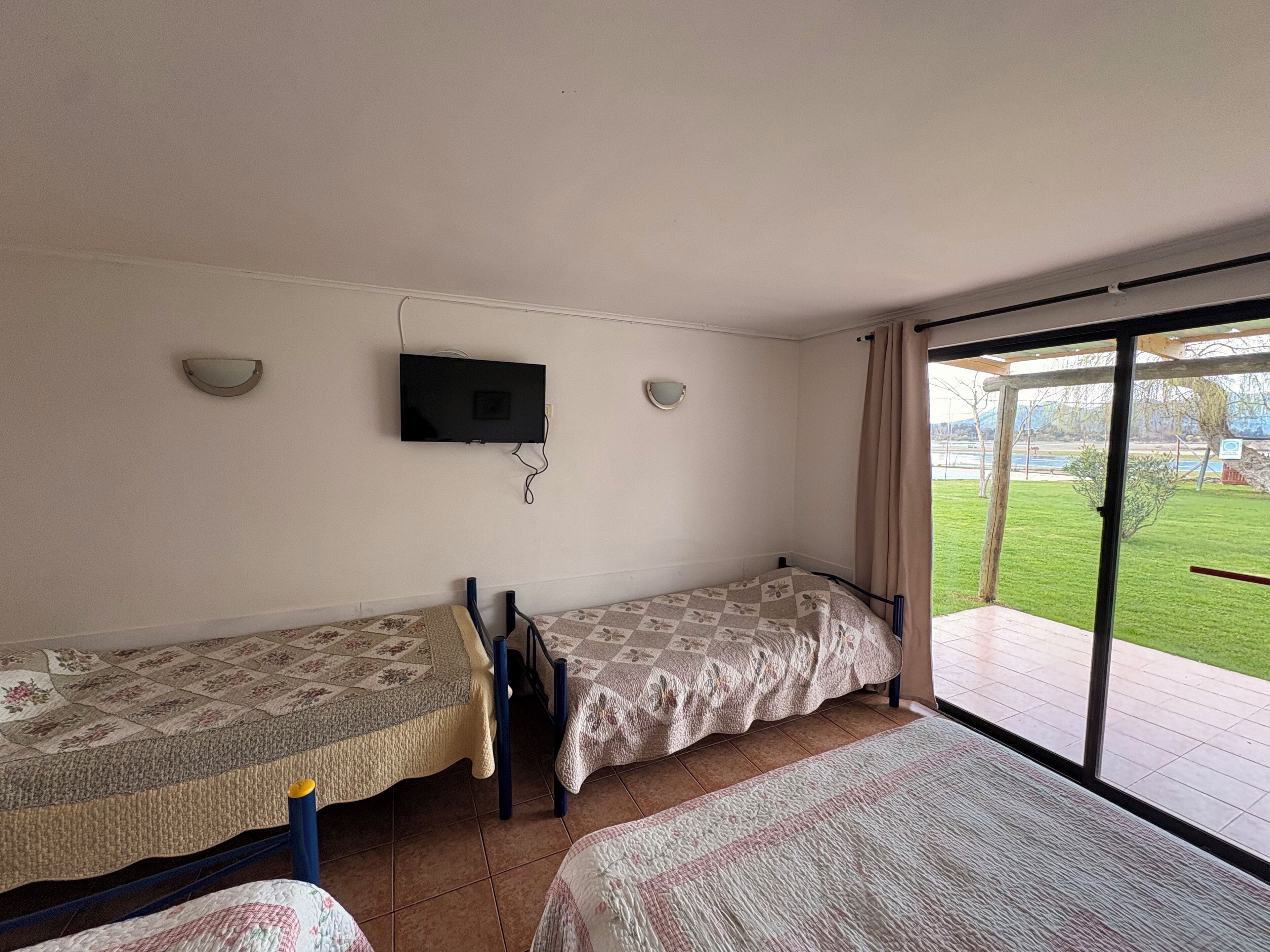 A room with two beds, a television mounted on the wall, and a sliding glass door leading to an outdoor area.