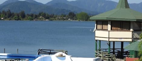 Vista de un lago con una pequeña torre de vigilancia, barcos y montañas al fondo.