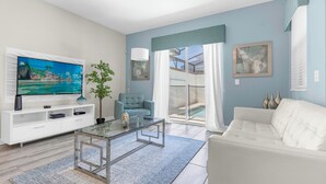 A modern living room with a flat-screen TV, a glass coffee table, a white sofa, and a sliding glass door leading to a balcony. - Champions Gate Resort Home with Pool (Four Corners)