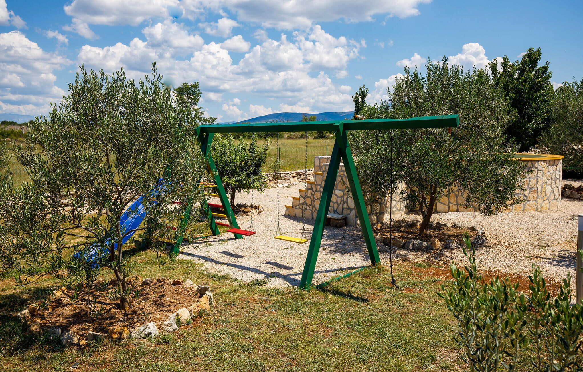 A playground with a swing set and climbing structures in a natural setting with trees and a stone wall.