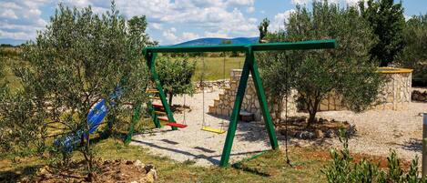 A playground with a swing set and climbing structures in a natural setting with trees and a stone wall.