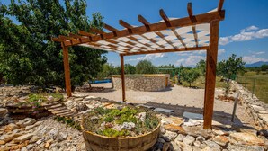 A wooden pergola structure with a stone bench underneath, surrounded by a gravel area with potted plants and a large wooden barrel filled with greenery.