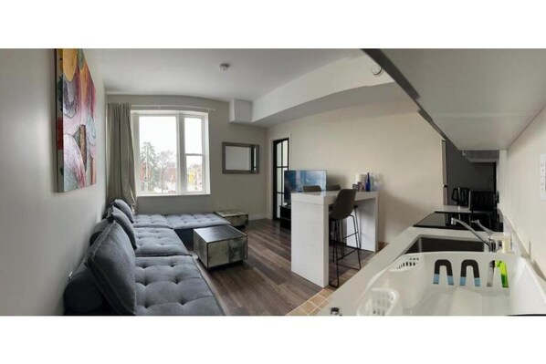 A modern living room with a grey sectional sofa, a small kitchen area with a white countertop, and a large window providing natural light.