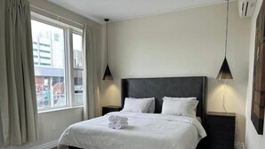 Quarto de hotel com uma cama grande, duas mesas de cabeceira, uma janela com cortinas e um aparelho de ar condicionado.