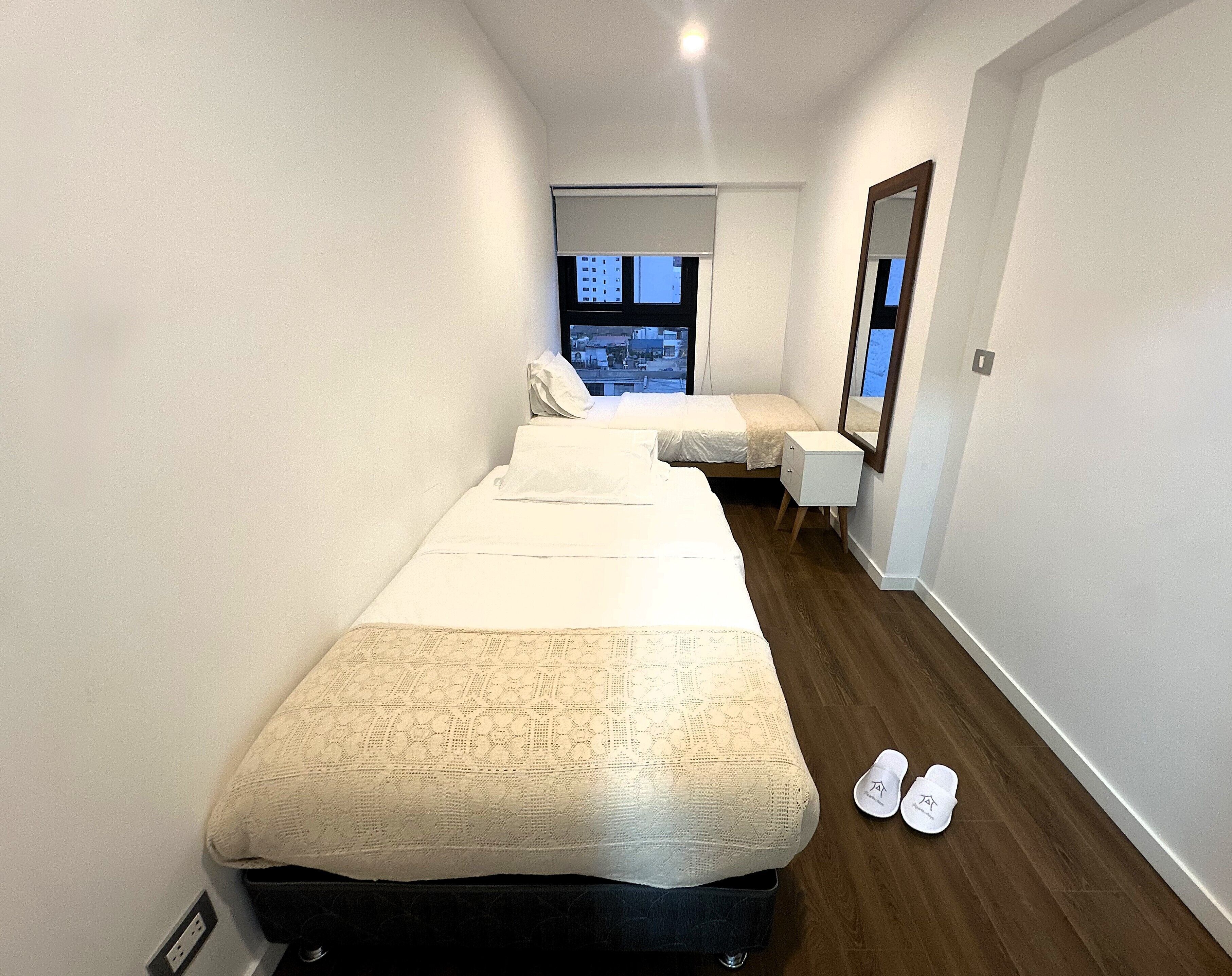 Quarto de hotel estreito com uma cama grande, uma cama menor, uma mesinha de cabeceira, um espelho e uma janela com vista.