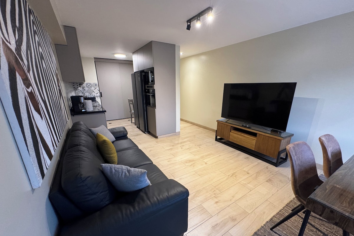 Una sala de estar moderna con un sofá de cuero negro, un televisor de pantalla plana sobre un soporte de madera, y un comedor con mesa y sillas.