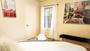 Habitación de hotel con cama, ventana con cortinas, una banca y un cartel en la pared con un autobús de dos pisos rojo.