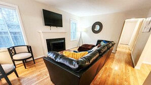 Una sala moderna con un sofá de cuero negro, una chimenea, un televisor montado en la pared y un área de comedor con una silla y una mesa.