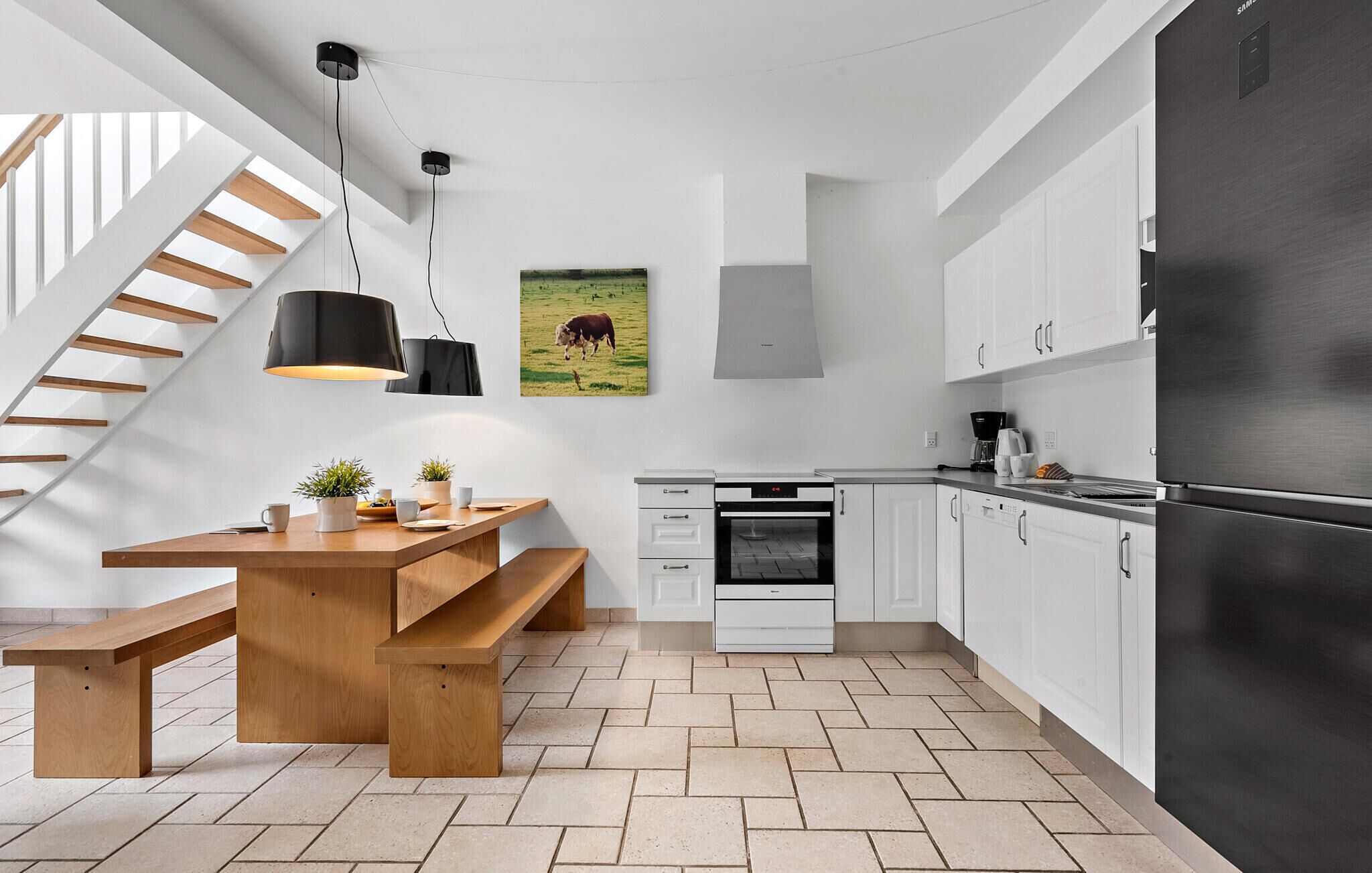 A modern kitchen with a wooden bench, pendant lights, a built-in oven, white cabinets, and a stainless steel refrigerator.