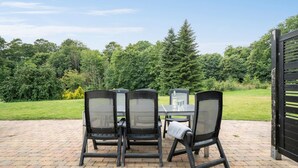 An outdoor patio with a table and four chairs, surrounded by a brick floor and a view of a forested area.