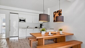 A modern kitchen with white cabinets, a built-in oven, and a coffee machine. There is a wooden dining table with benches and two pendant lights.