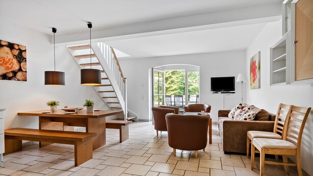 A modern living room with a wooden staircase, a dining area, and a large window offering a view of greenery.