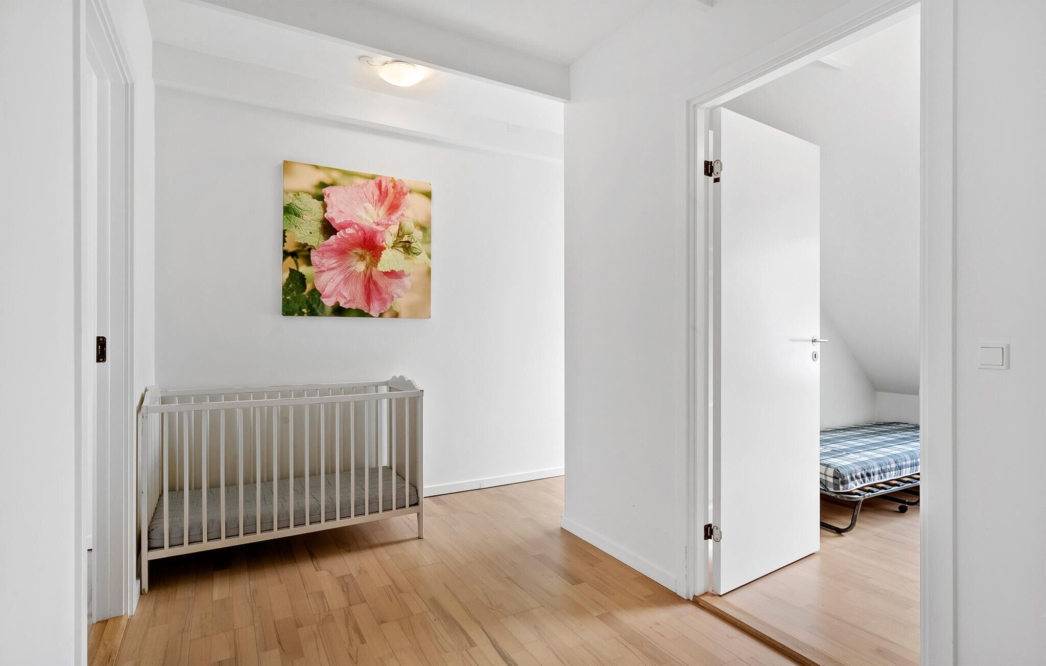 A room with a white crib, a bed with a checkered blanket, and a door leading to another room.