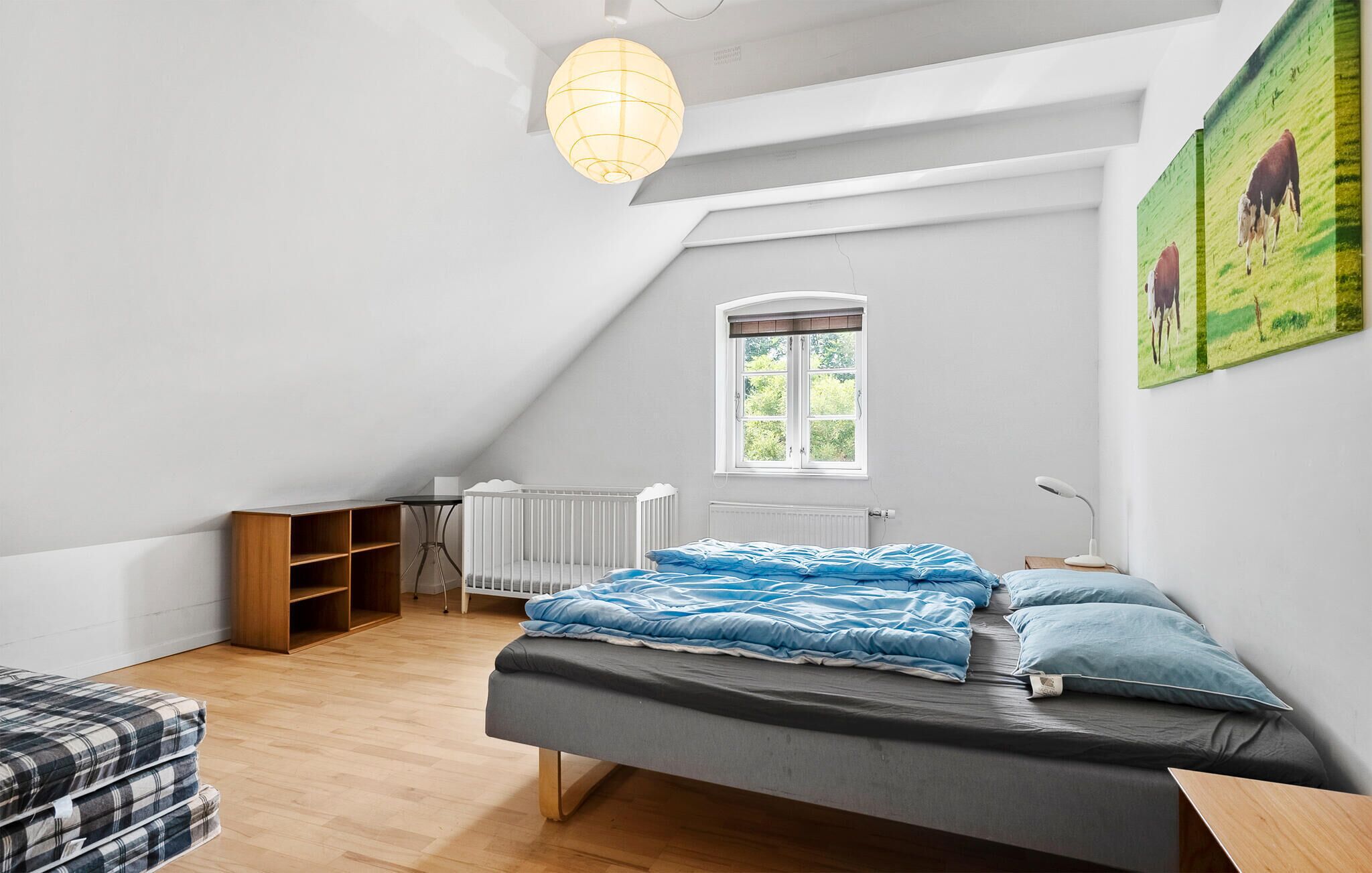 Ett sovrum med ett sluttande tak, ett fönster med utsikt över grönområden, en spjälsäng, ett nattduksbord med en lampa och en tavla med kor på väggen.