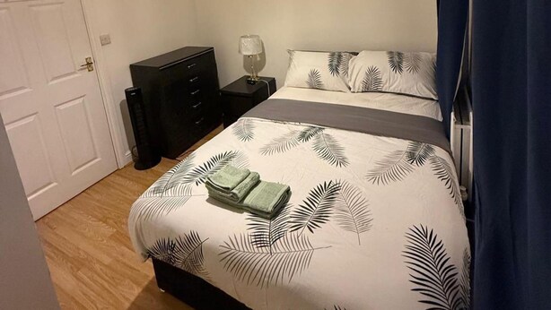 Ein ordentlich bezogenes Bett mit einem blattgemusterten Überwurf, ein kleiner Nachttisch mit einer Lampe und eine dunkle Kommode.